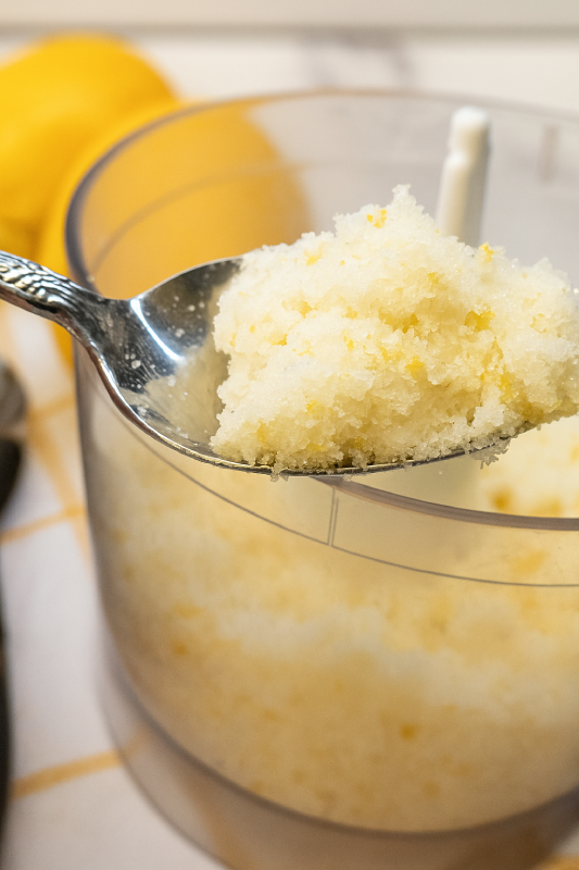Lemon Zest Sugar Trick A close-up of a spoonful of lemon sugar held above a food processor bowl filled with the citrus-infused mixture.