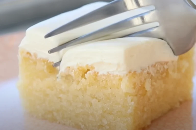 A fork presses into a moist slice of vanilla cake topped with creamy white frosting.